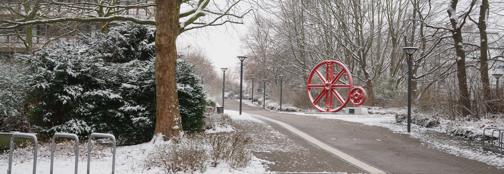 Ein verschneiter Gehweg zu den rot-lackierten Zahnrädern auf dem Campus Nord.